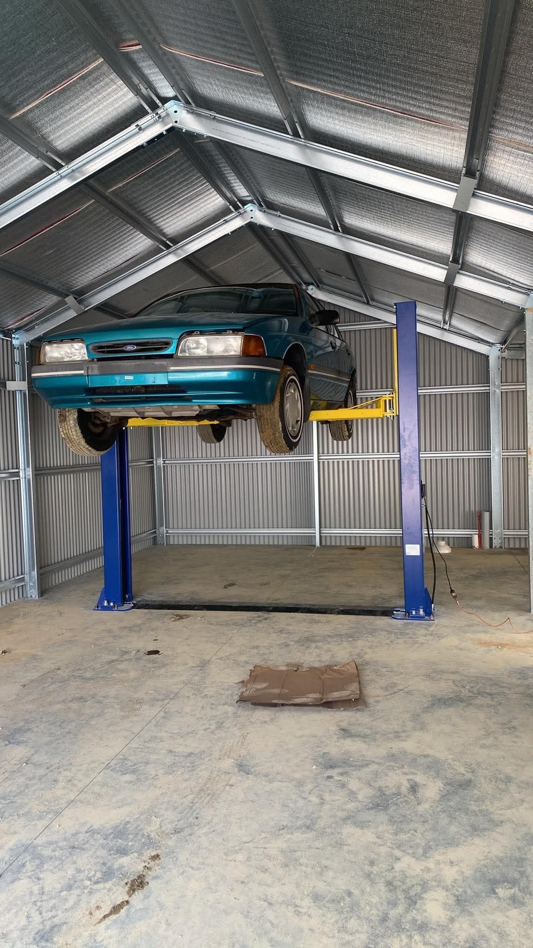 Turquoise car lifted on a blue hydraulic lift inside a metal garage shed