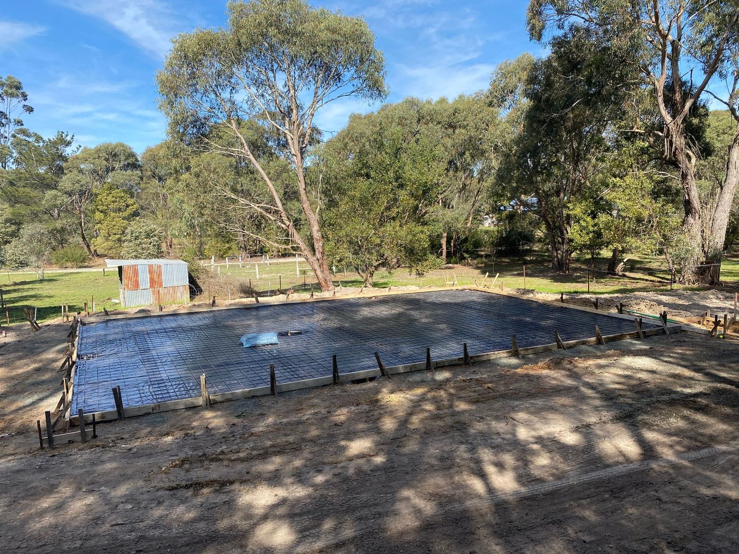 Outdoor construction site with a square concrete slab, rebar stakes, and trees in the background.