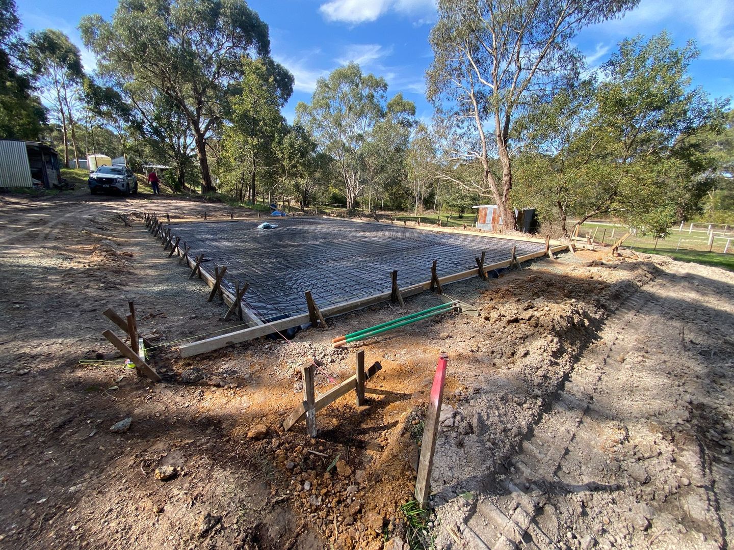 Concrete foundation under construction on a dirt lot with trees and exposed plumbing pipes