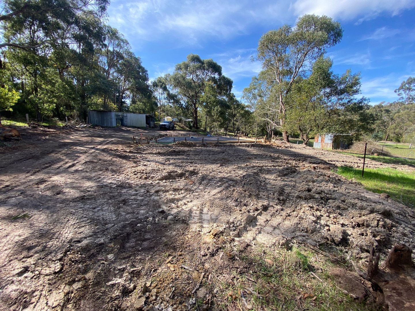 Cleared dirt lot under trees beside a fence, with a wooded hillside in the background