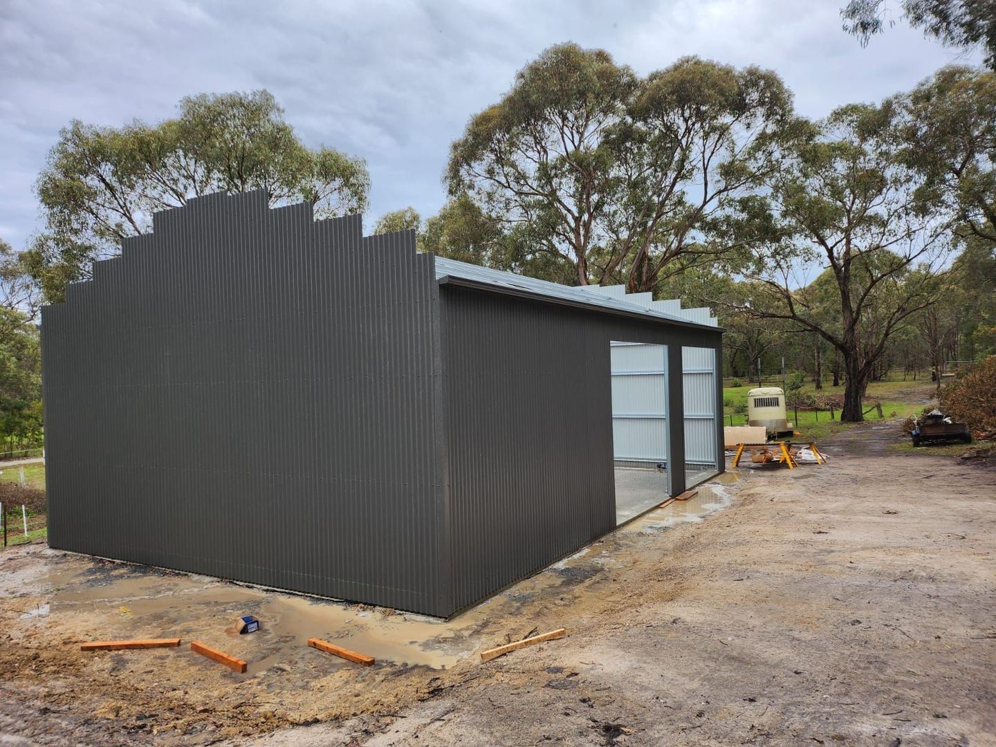 Small dark corrugated shed with glass door on a dirt site, surrounded by trees and construction materials.