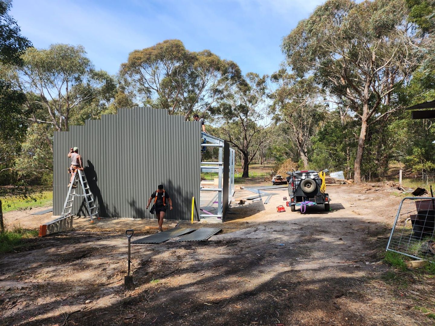 Corrugated metal shed under construction with tools, a ladder, and equipment on a gravel site surrounded by trees