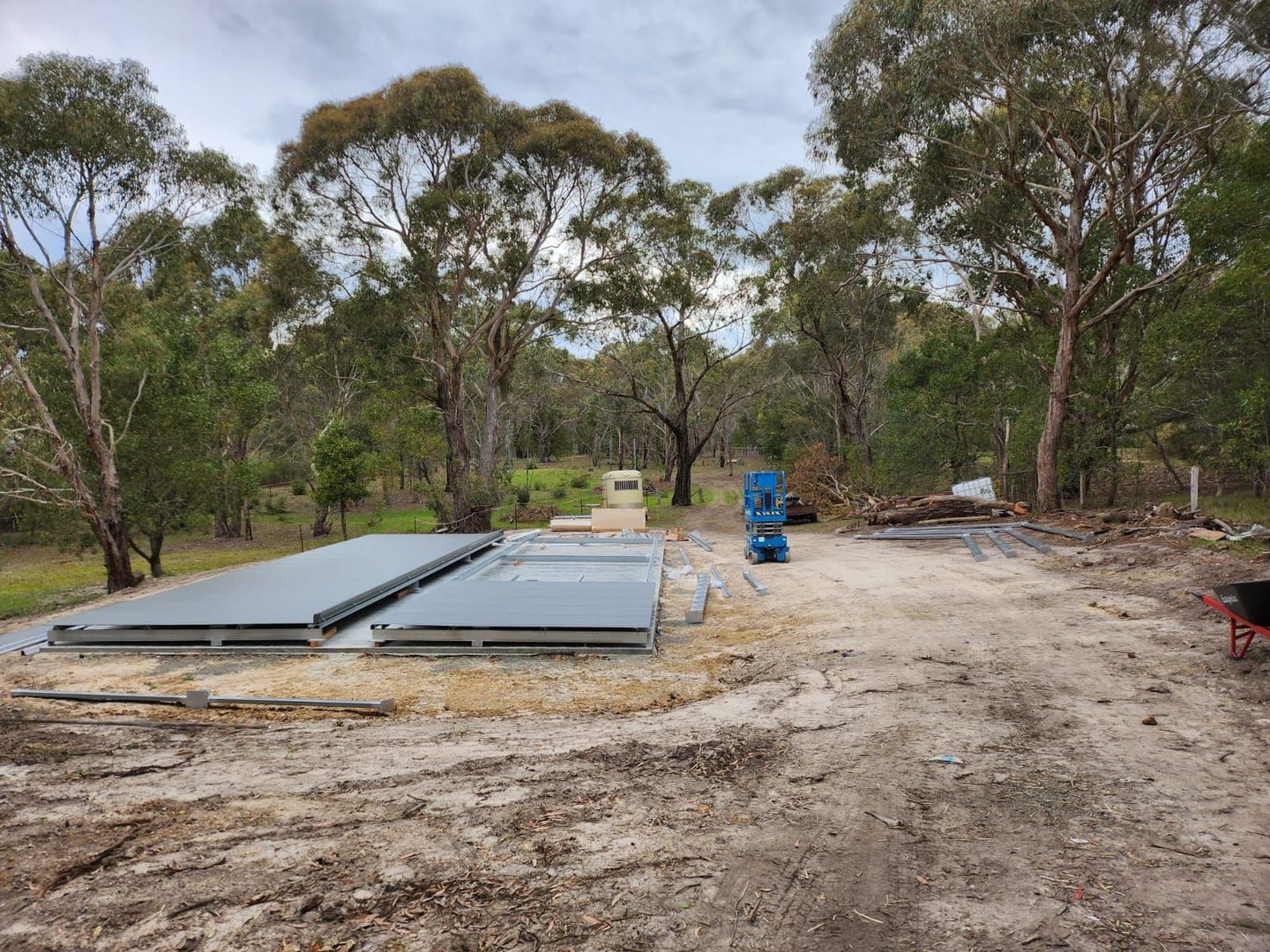 Rural construction site with stacked steel panels, a blue machine, and trees along a dirt road