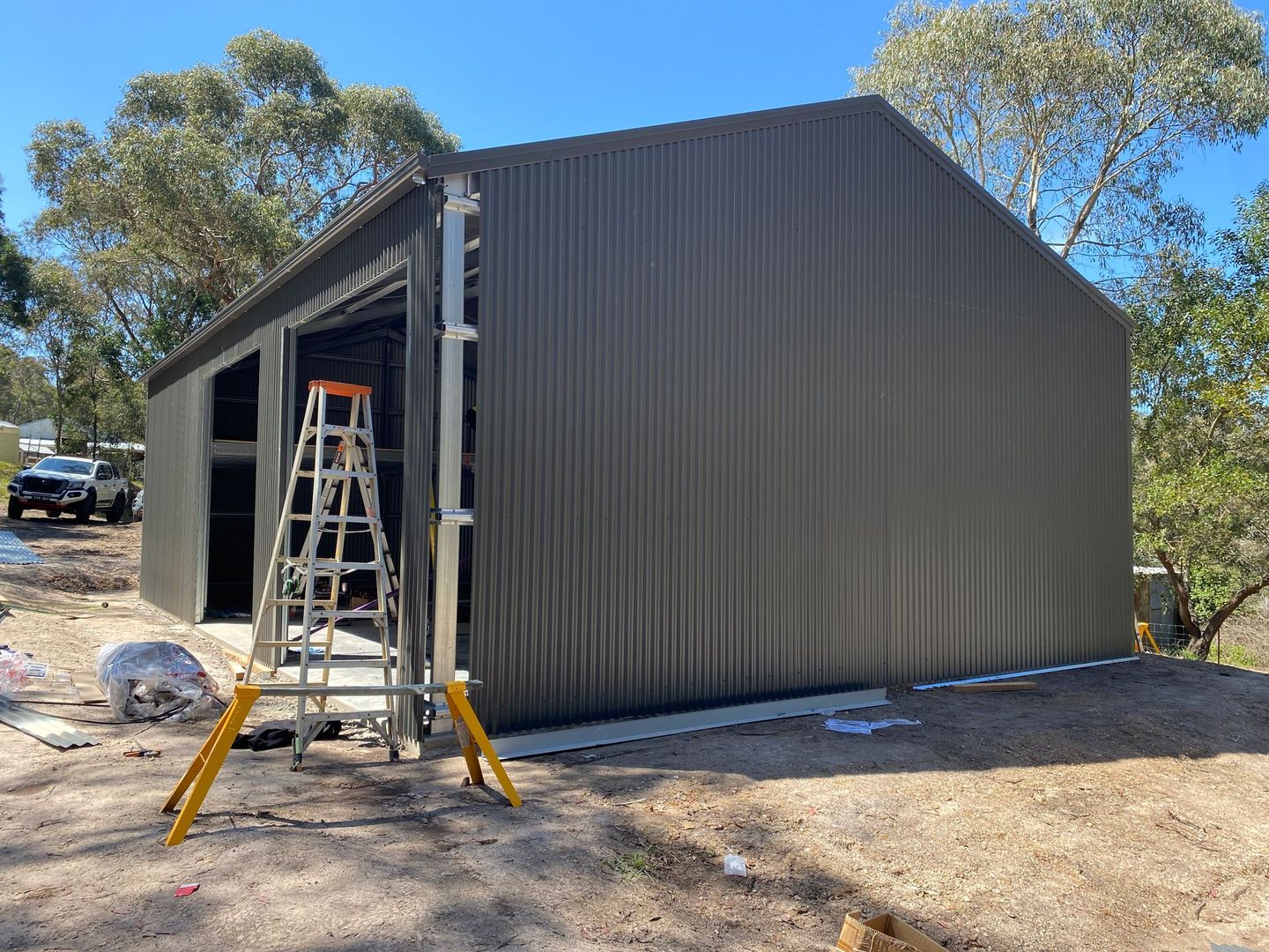 Gray metal shed under construction with open doorway, ladder, and yellow sawhorses outdoors in a wooded area