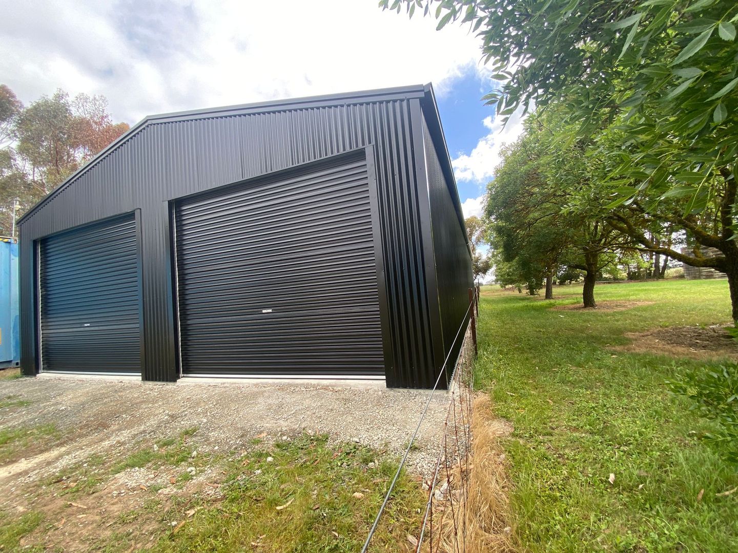 Black metal shed with roller doors on a grassy roadside under trees and a cloudy sky