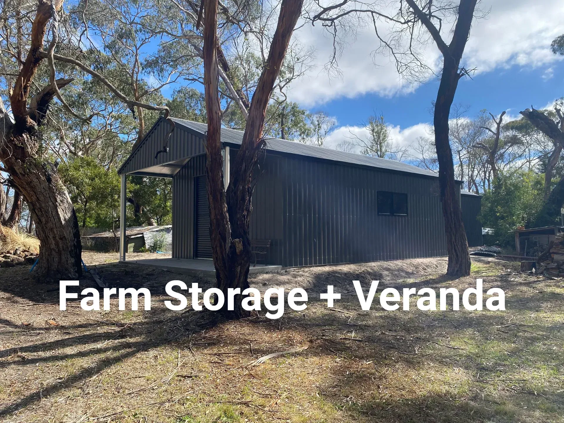 Dark metal farm storage shed with a veranda, surrounded by trees under a blue sky.