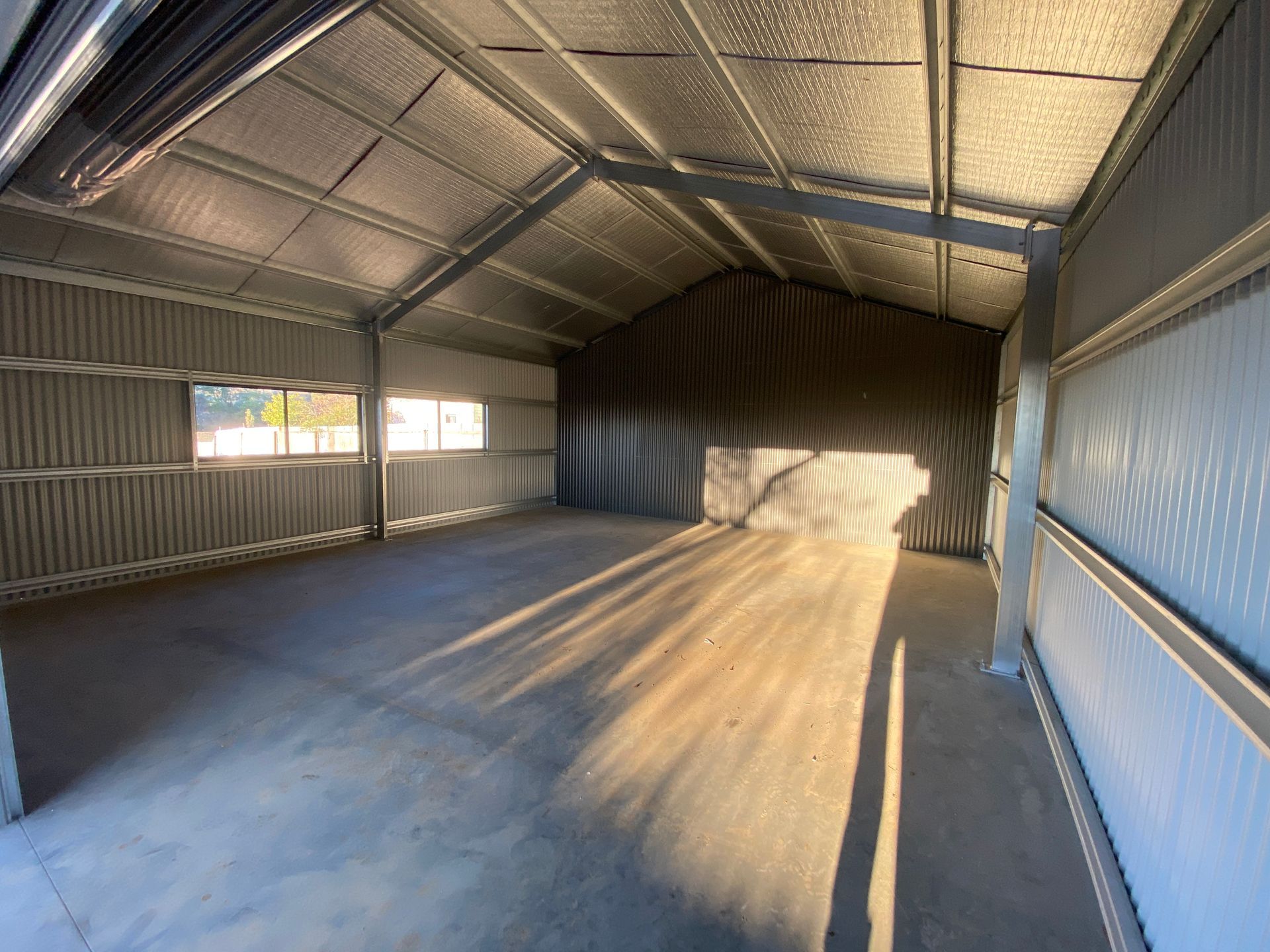 Empty metal storage shed with corrugated walls, concrete floor, and sunlight streaming through windows.