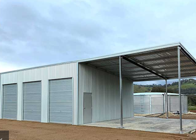 Metal storage shed with roll-up doors and an open-sided carport on a dirt lot