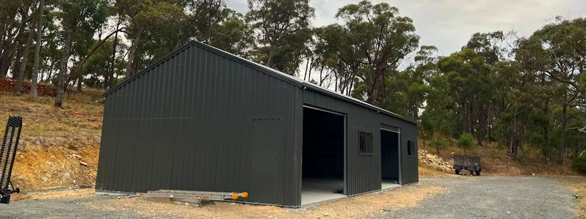 Dark gray metal shed with open doorway and windows, set on a dirt lot with trees behind.