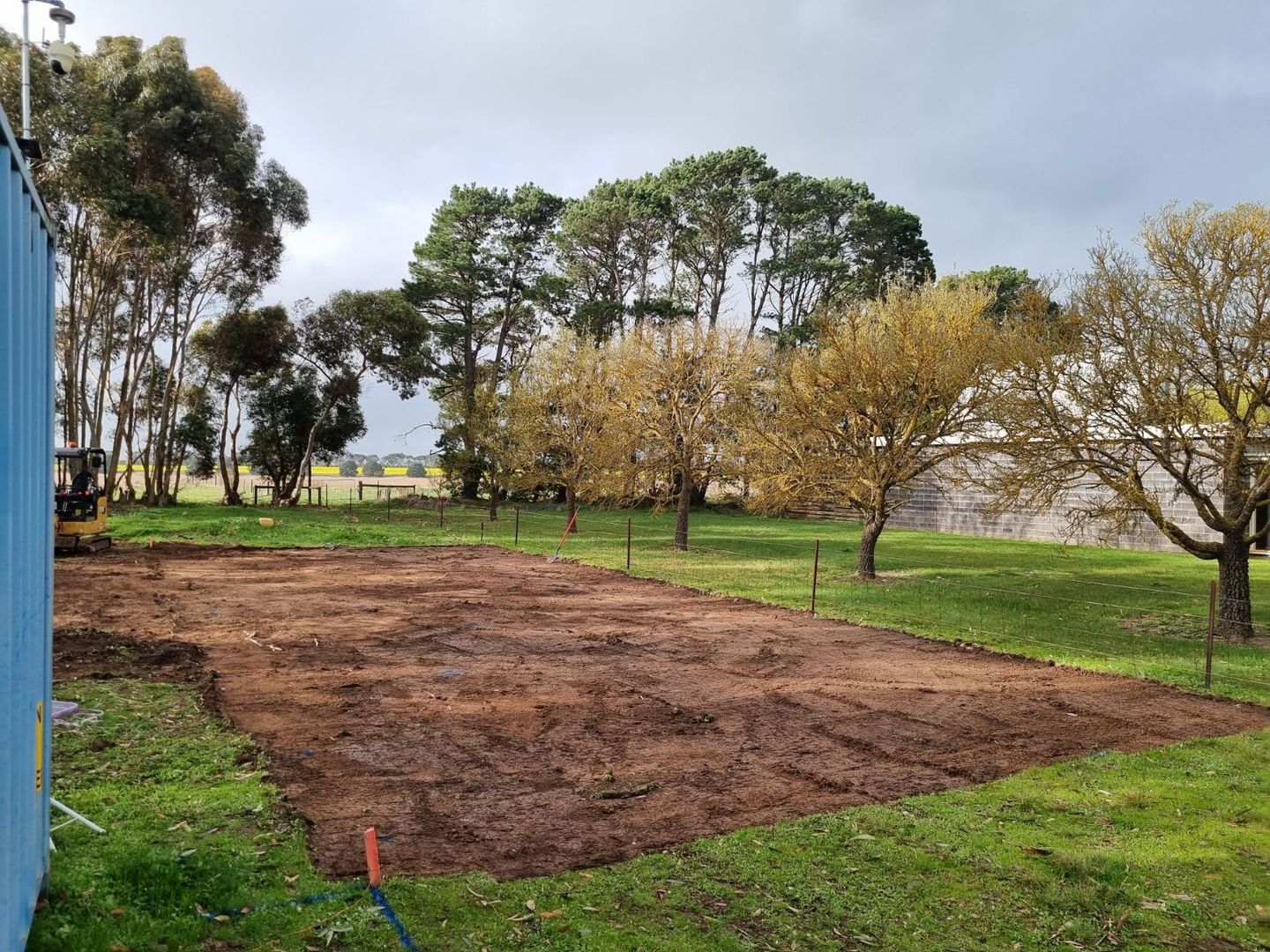 Freshly tilled brown soil patch in a grassy park with trees and cloudy sky