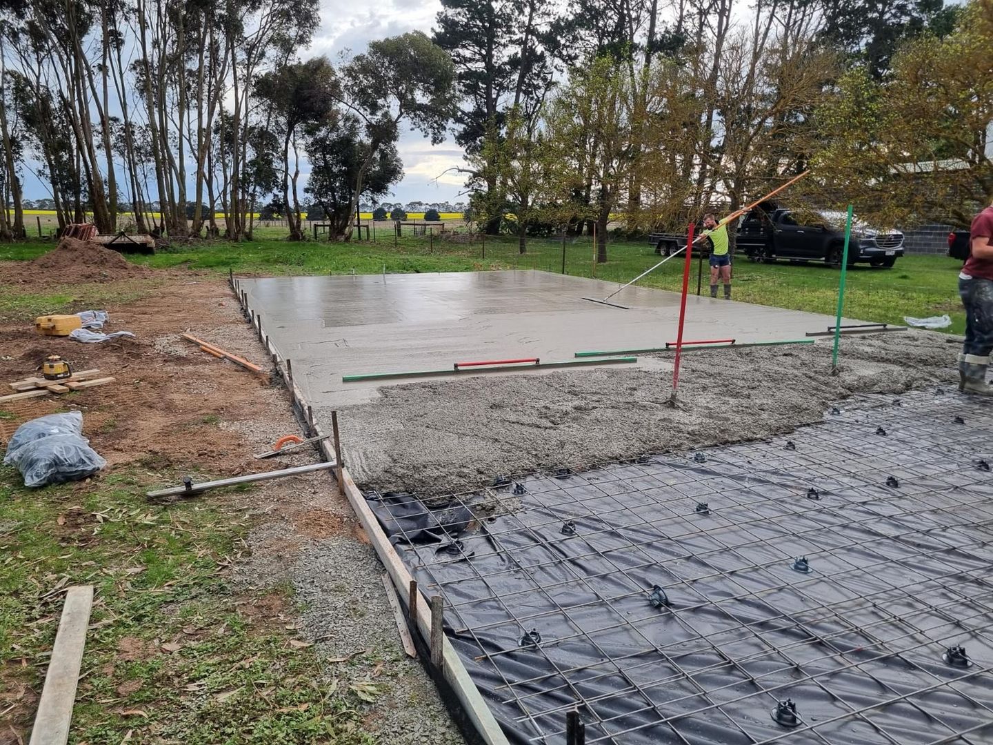 Concrete slab and gravel work area at a park, with a worker and trees in the background.