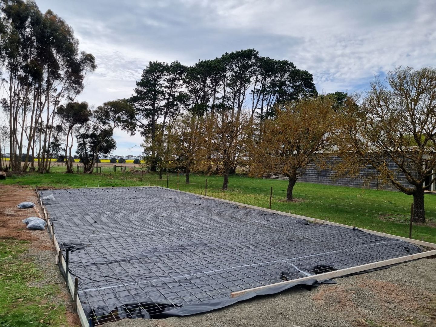 Large gray-covered rectangular plot in a grassy park with trees and cloudy sky