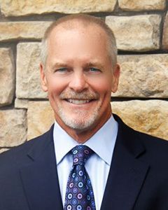 A man in a suit and tie is smiling in front of a stone wall.