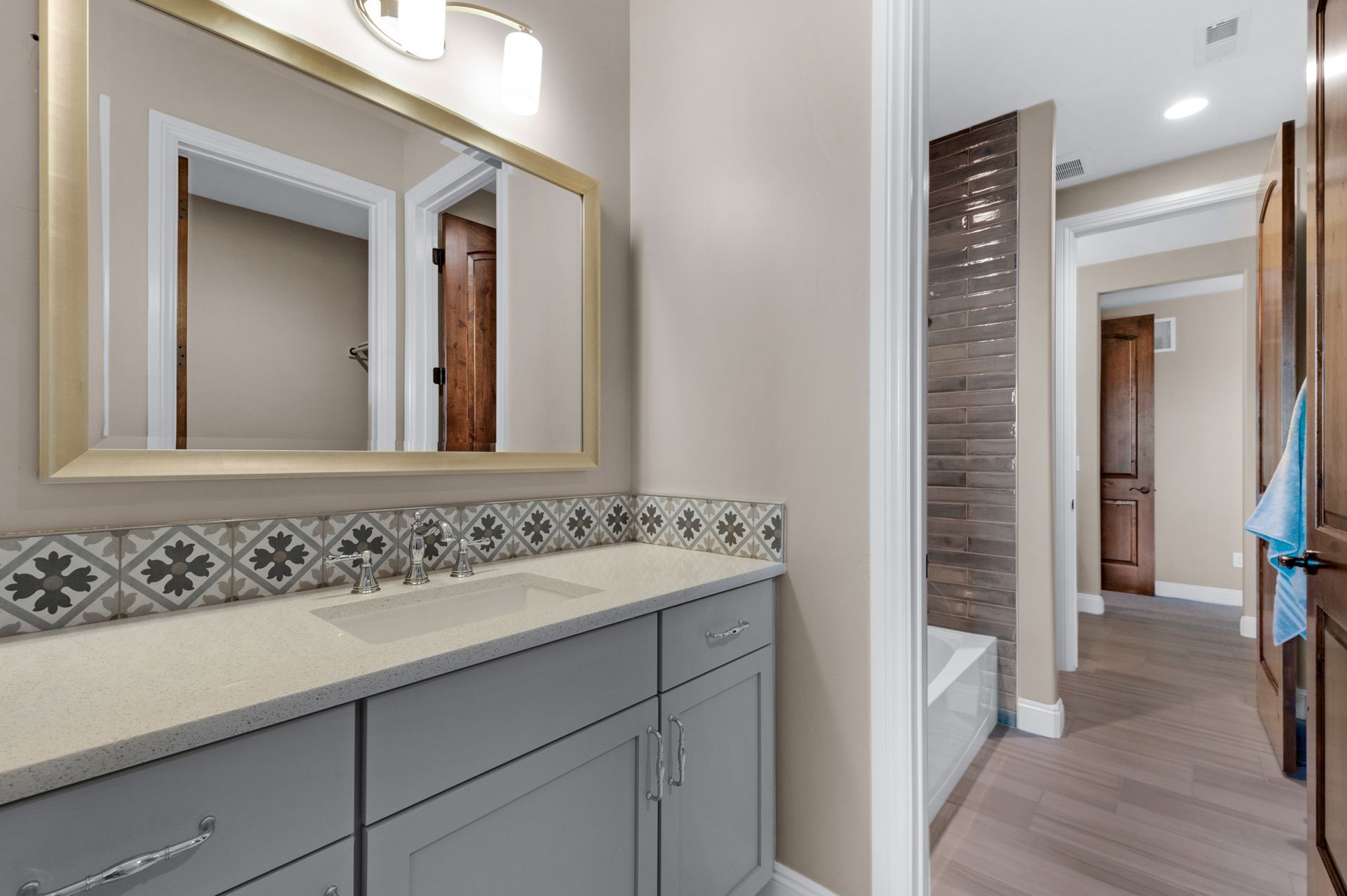Gray vanity in bathroom with patterned backsplash, large mirror. Hallway visible.