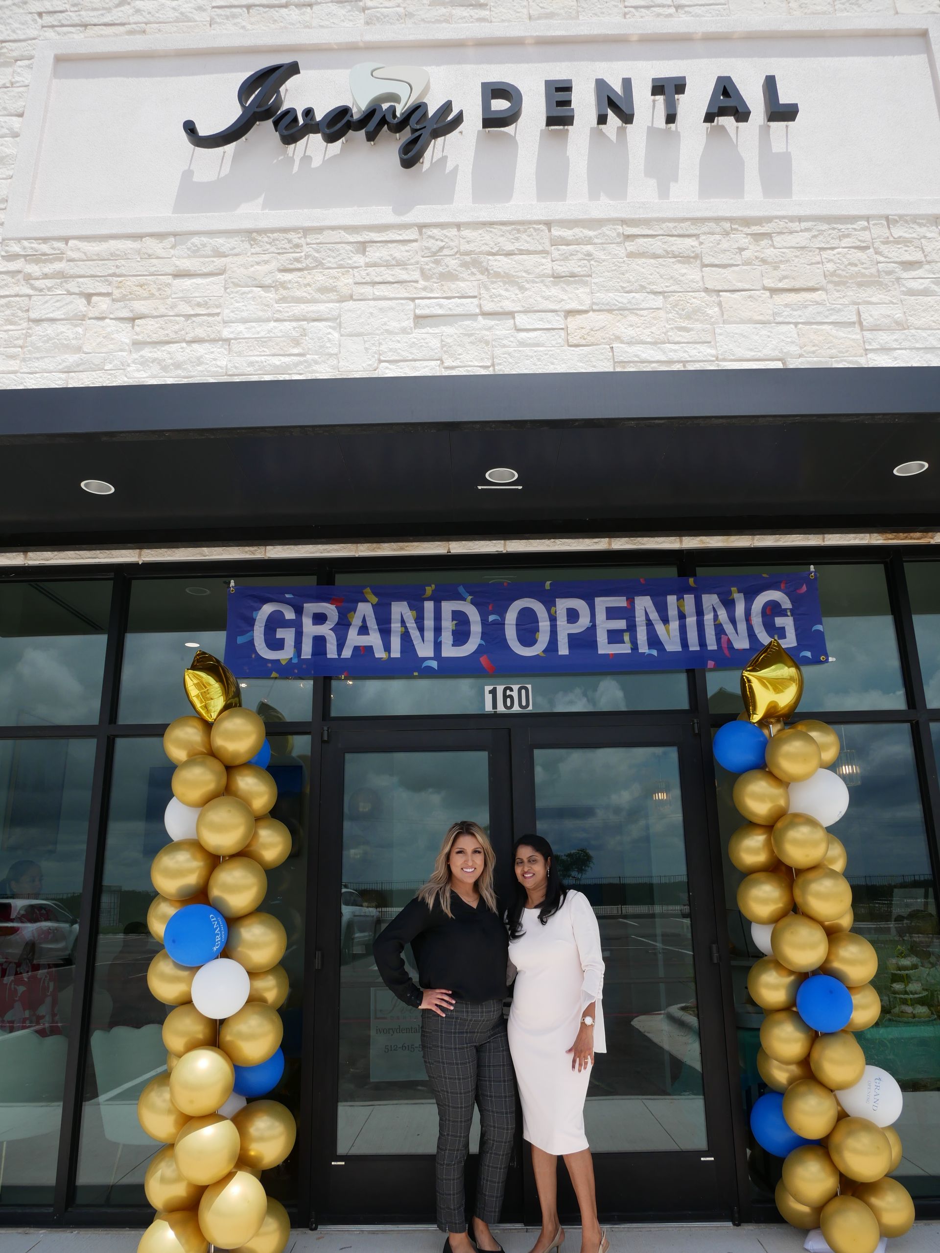 Two women are standing in front of a building with balloons and a sign that says grand opening.