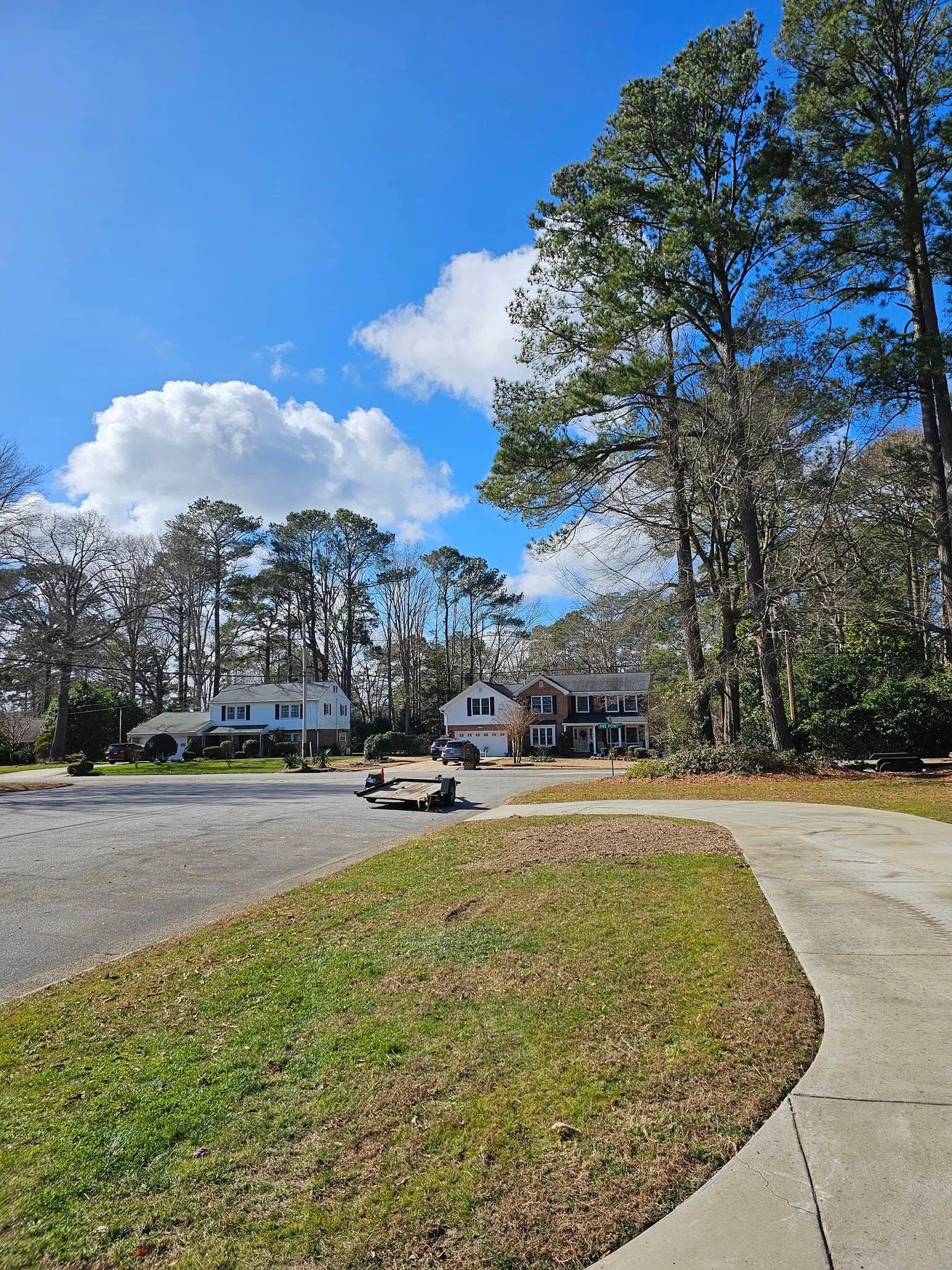 Curved sidewalk beside a grassy median under blue sky, with trees and houses in the distance.