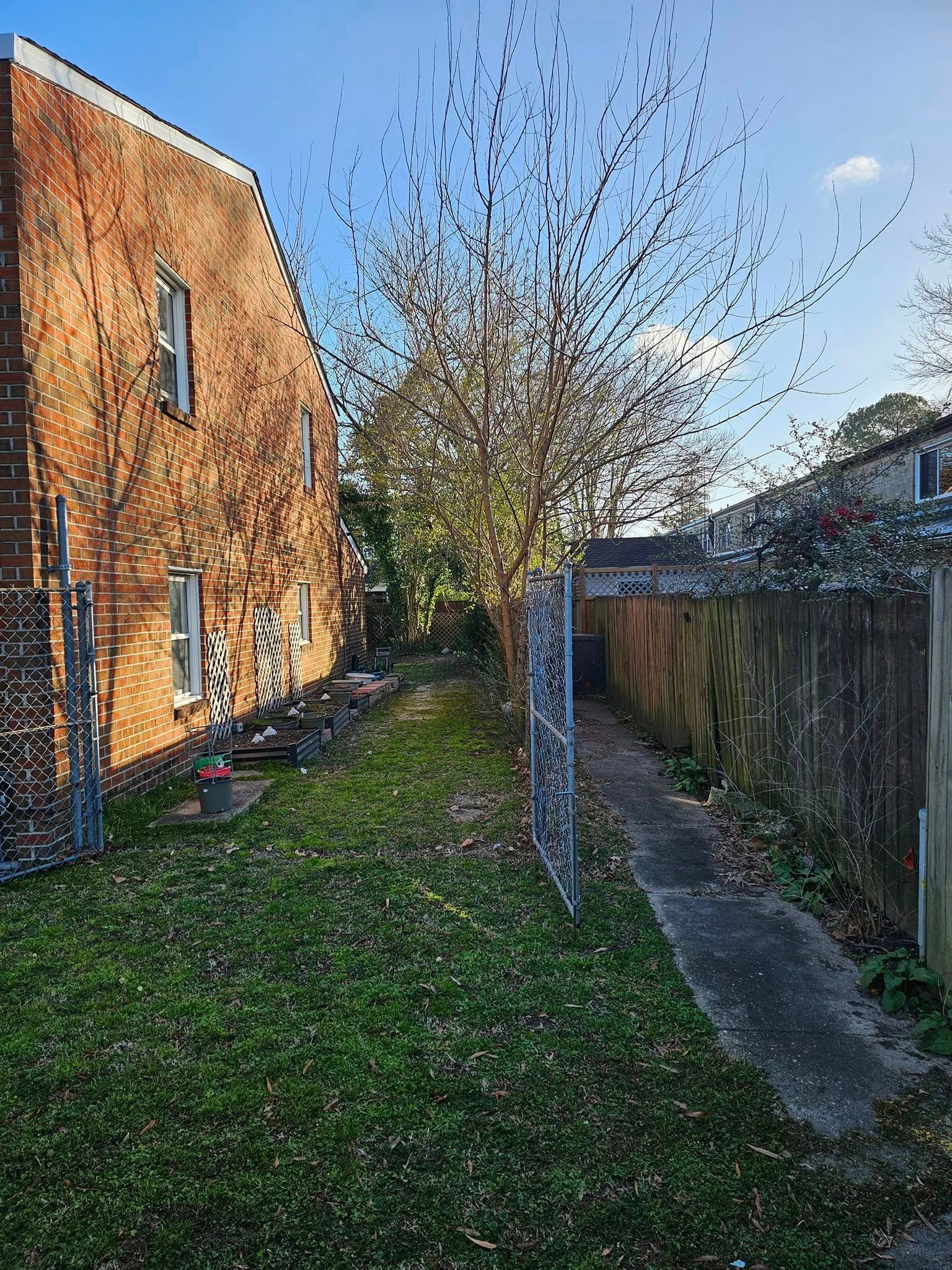 Narrow backyard with a brick house, grassy lawn, leafless tree, and a concrete path beside a wooden fence.