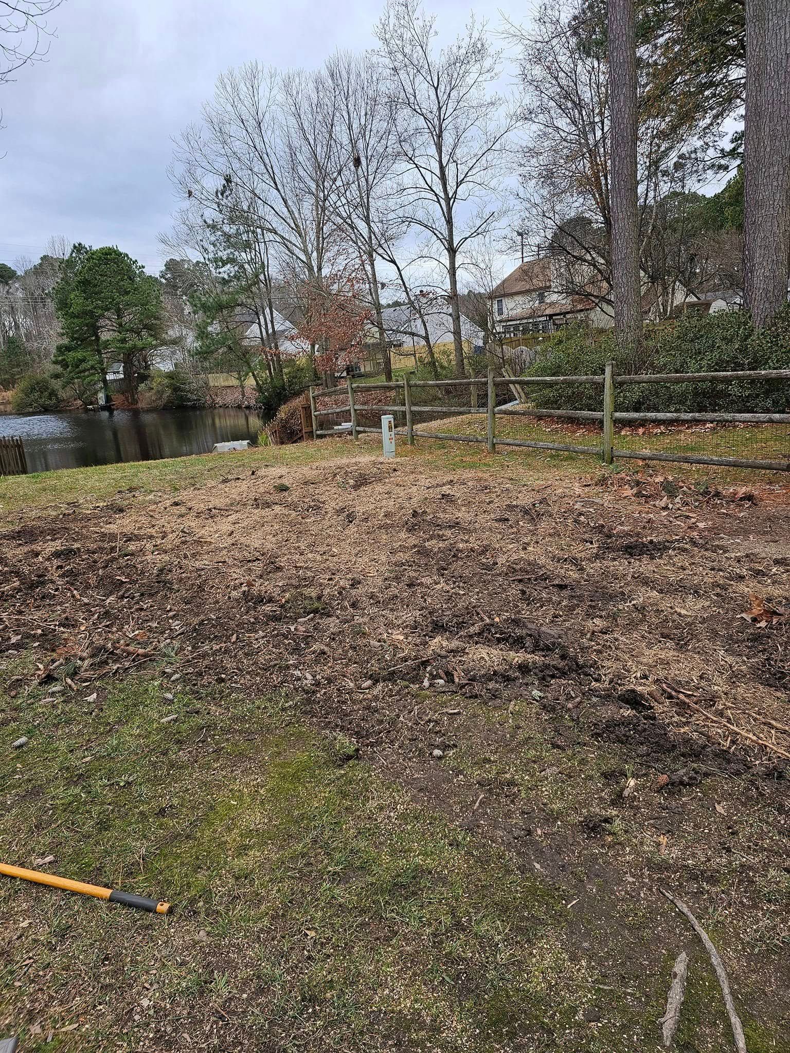 Leaf-covered yard beside a calm pond, with bare trees and a fence in the background.