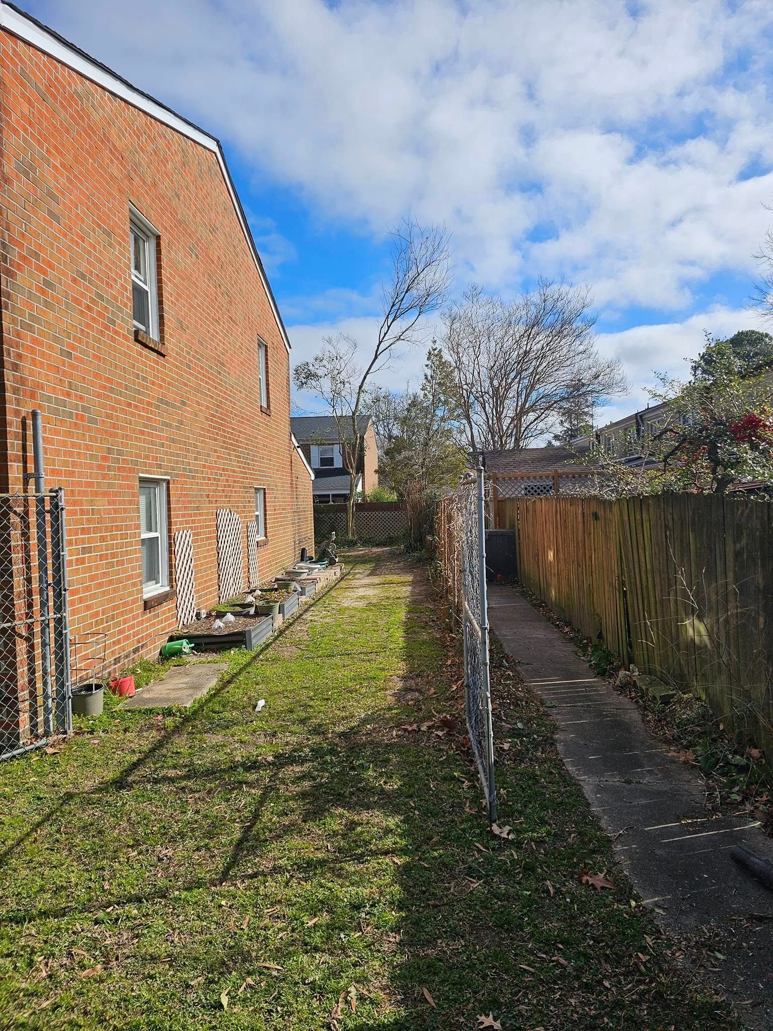 Backyard beside a brick house with a narrow grassy path, fence, and a young tree under a cloudy sky