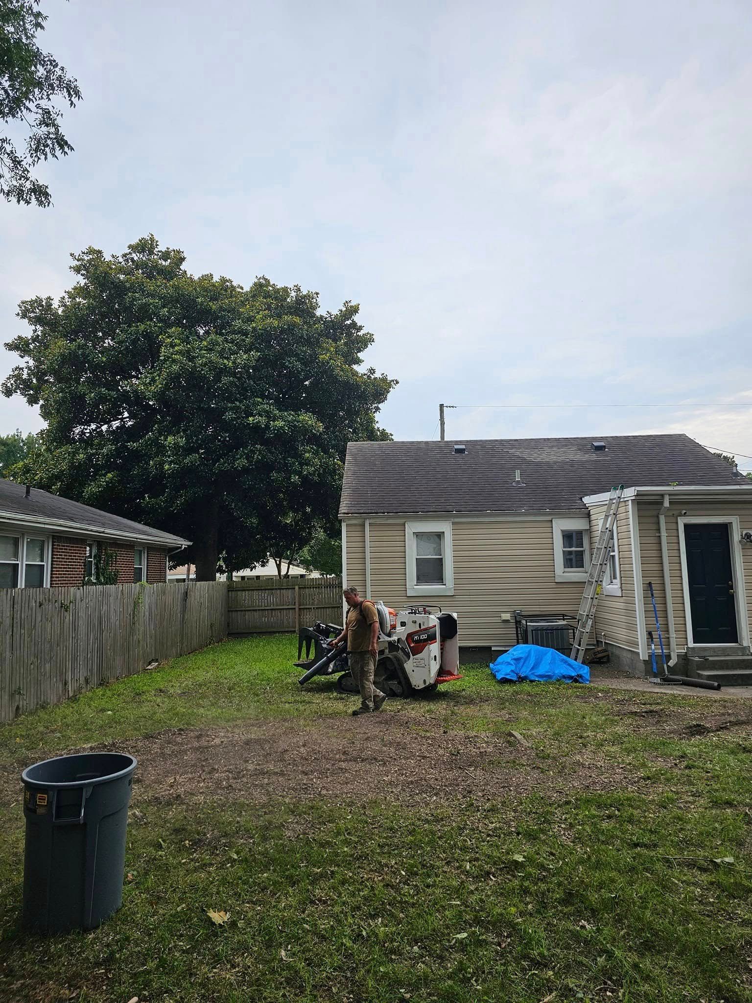 Backyard with a beige house, tree, fence, lawn, trash bin, and a motorcycle under a cloudy sky.