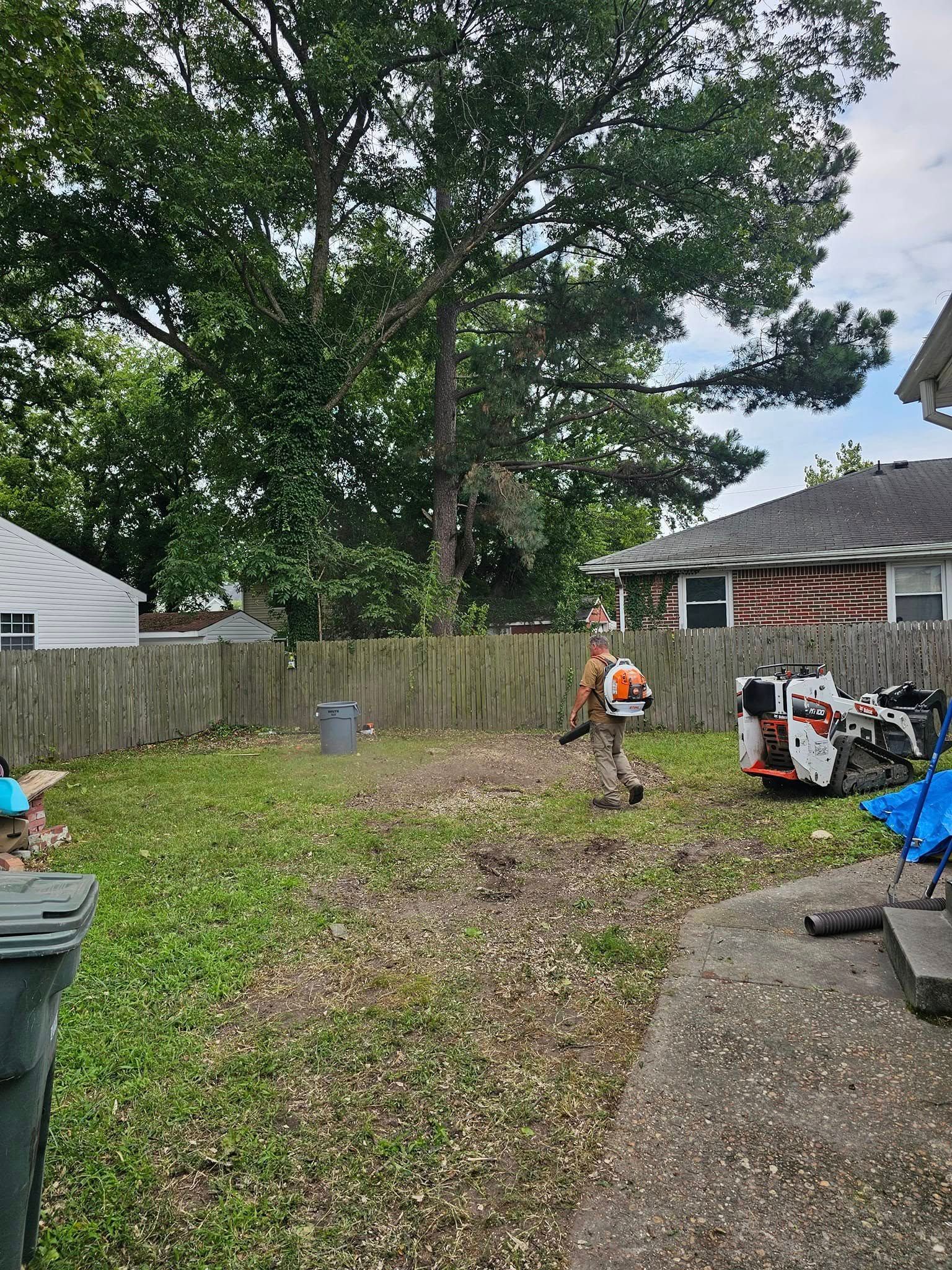 Backyard with a large tree, wooden fence, and a person in an orange vest clearing leaves and debris