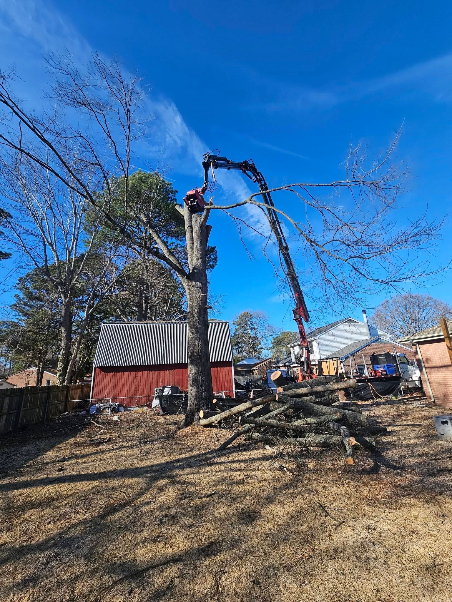Excavator lifting debris beside a red barn under a blue sky, with a tree and utility truck nearby