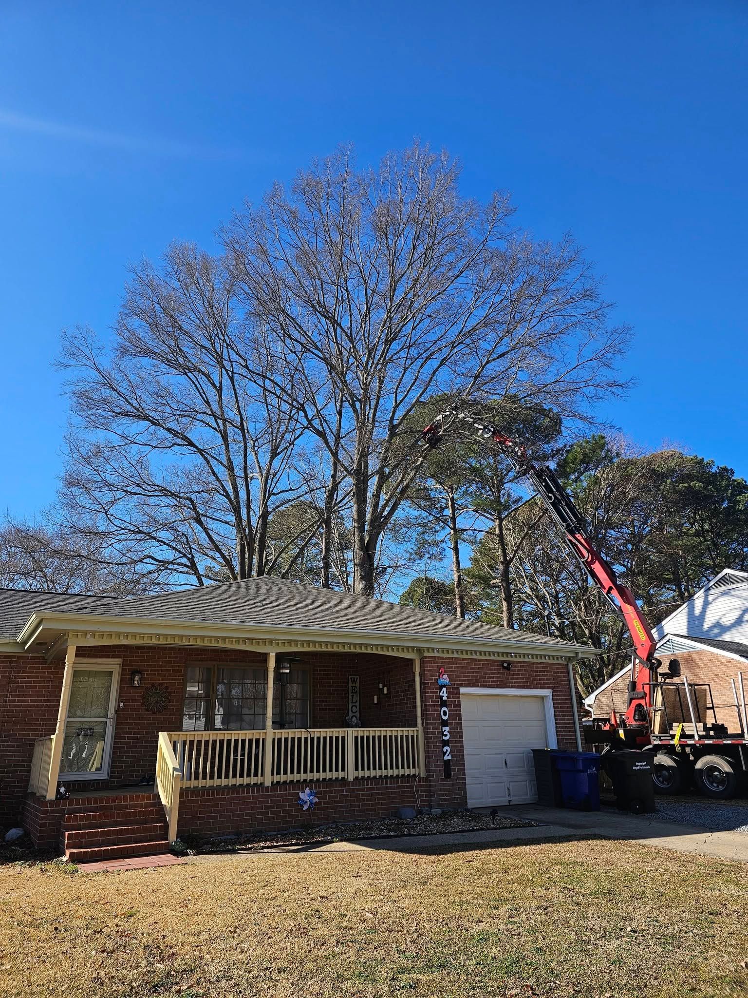 Single-story brick house with a porch, large leafless tree, and a red crane truck in the yard under a blue sky