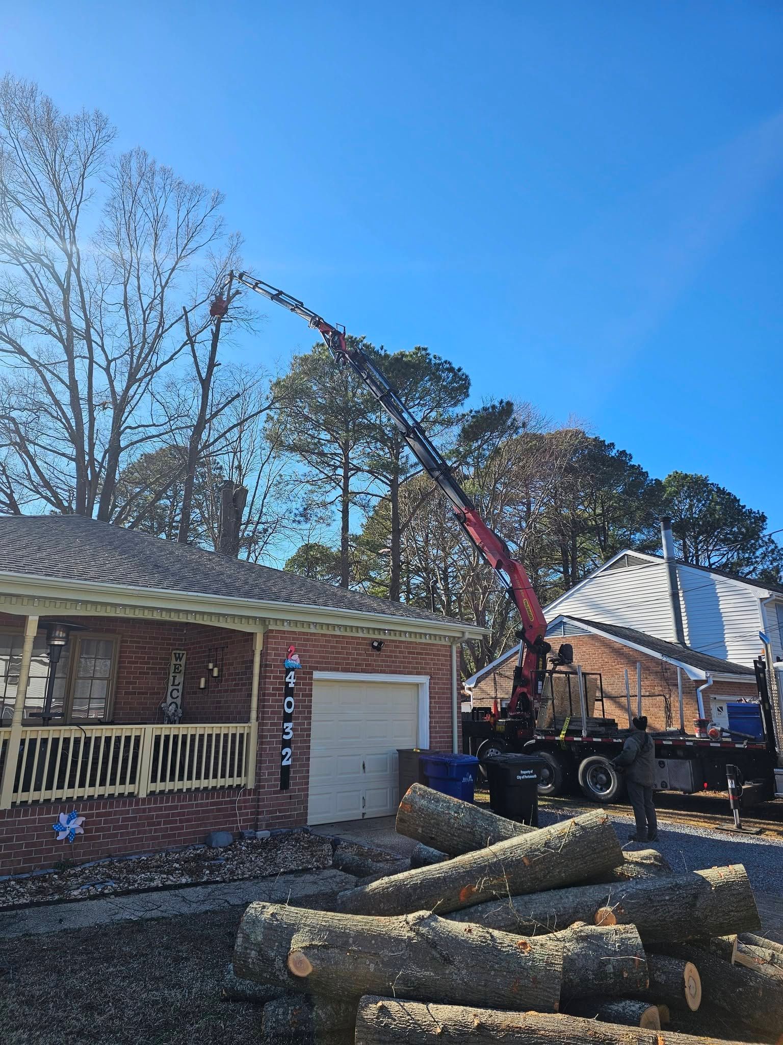 Crane lifting tree limbs over a brick house roof, with cut logs stacked in the foreground.