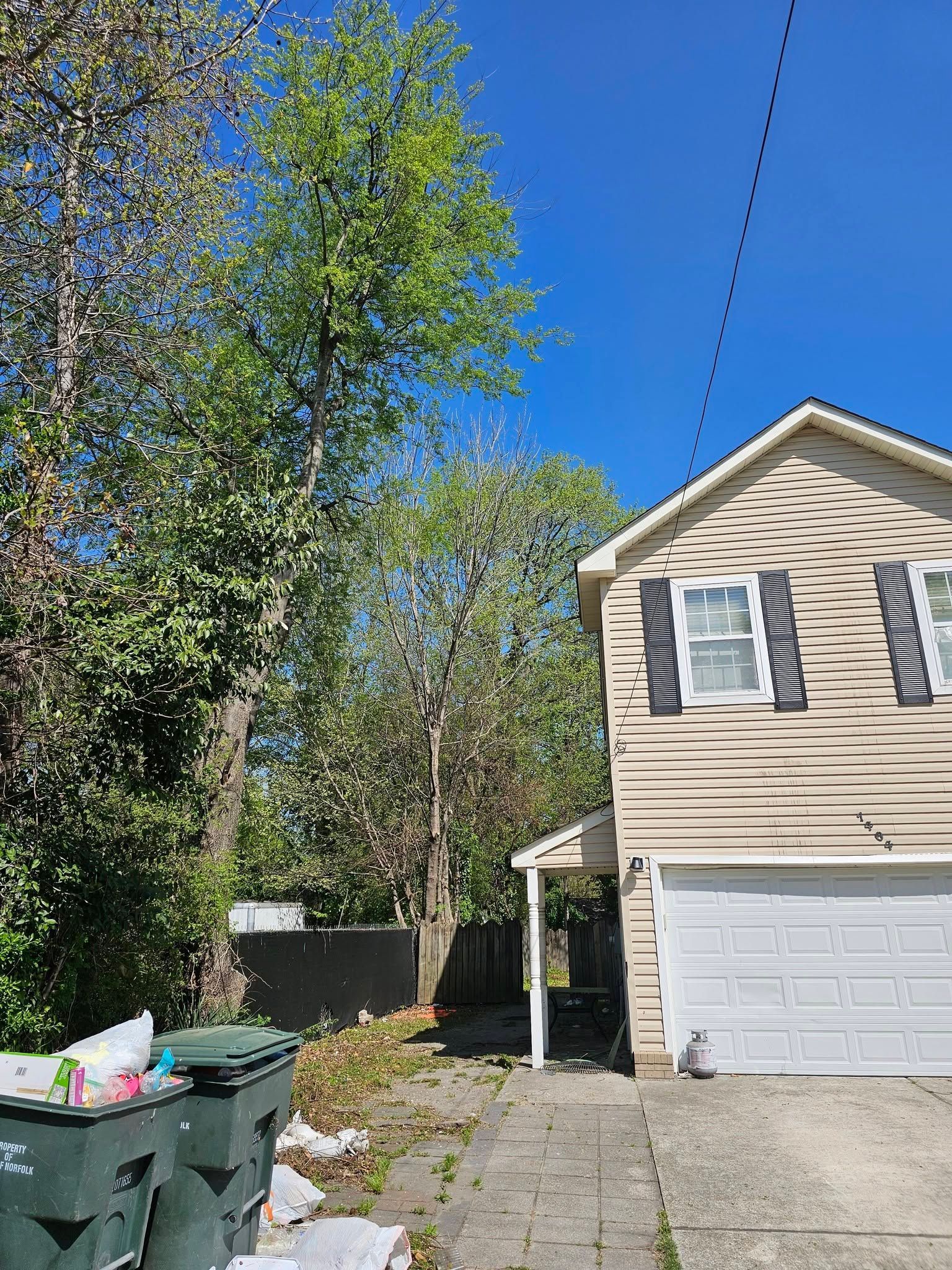 Two-story beige house with garage, driveway, trash bins, and trees under a clear blue sky