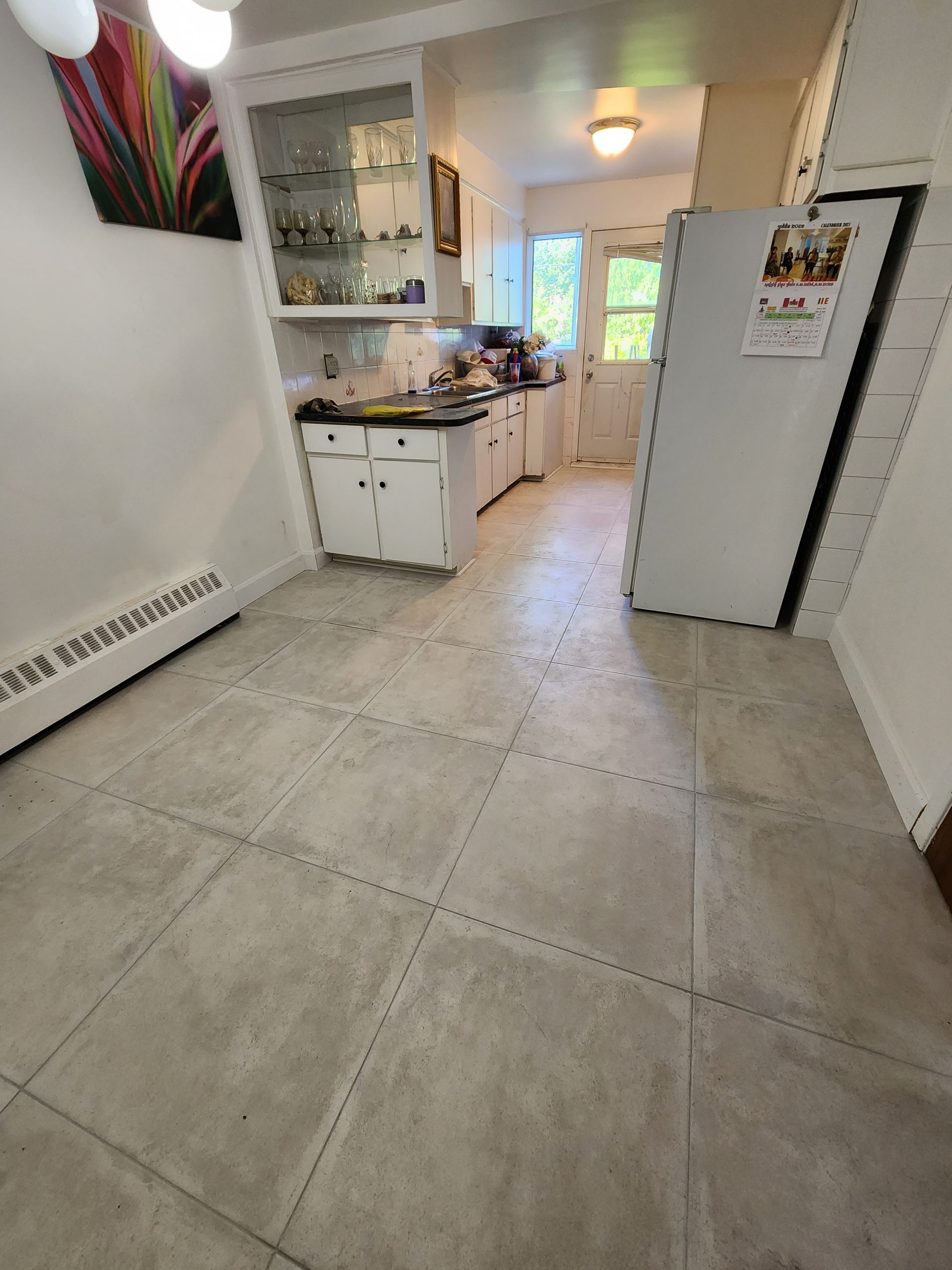 A kitchen with tile floors , white cabinets , a refrigerator and a painting on the wall.