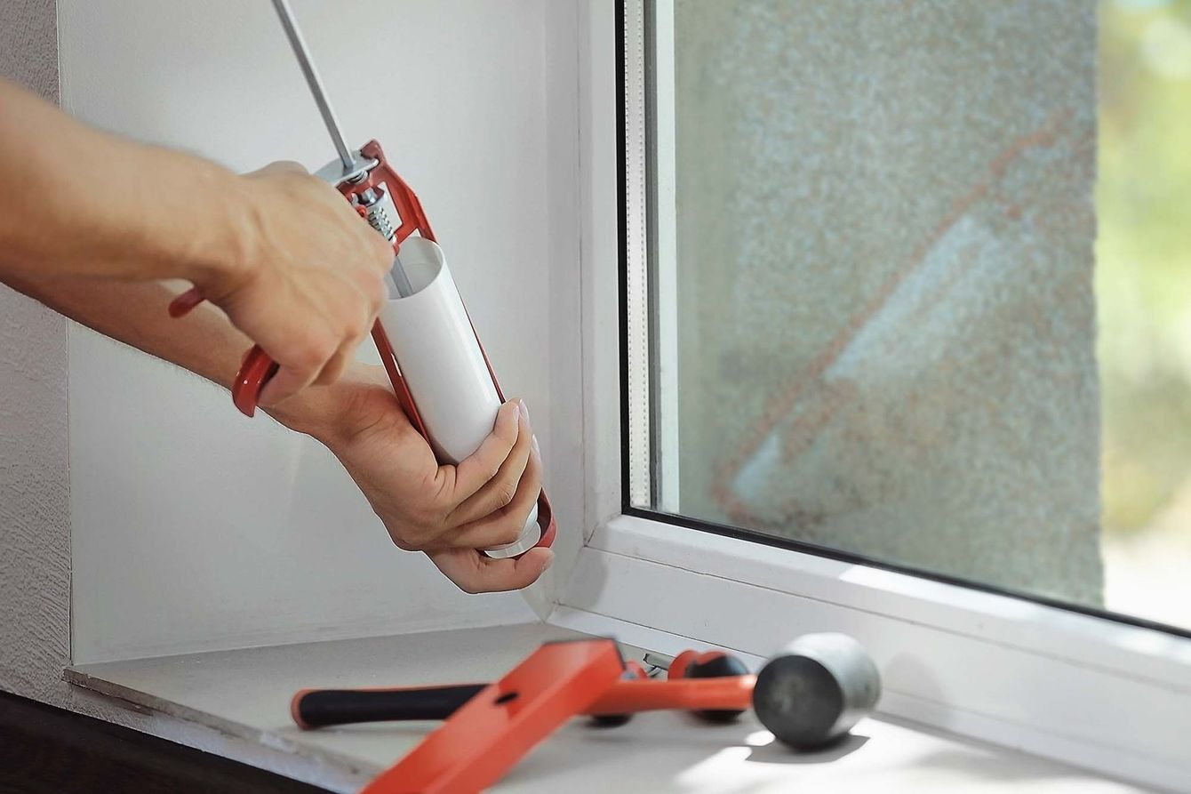 Hands applying caulk from a red caulking gun to seal a window frame