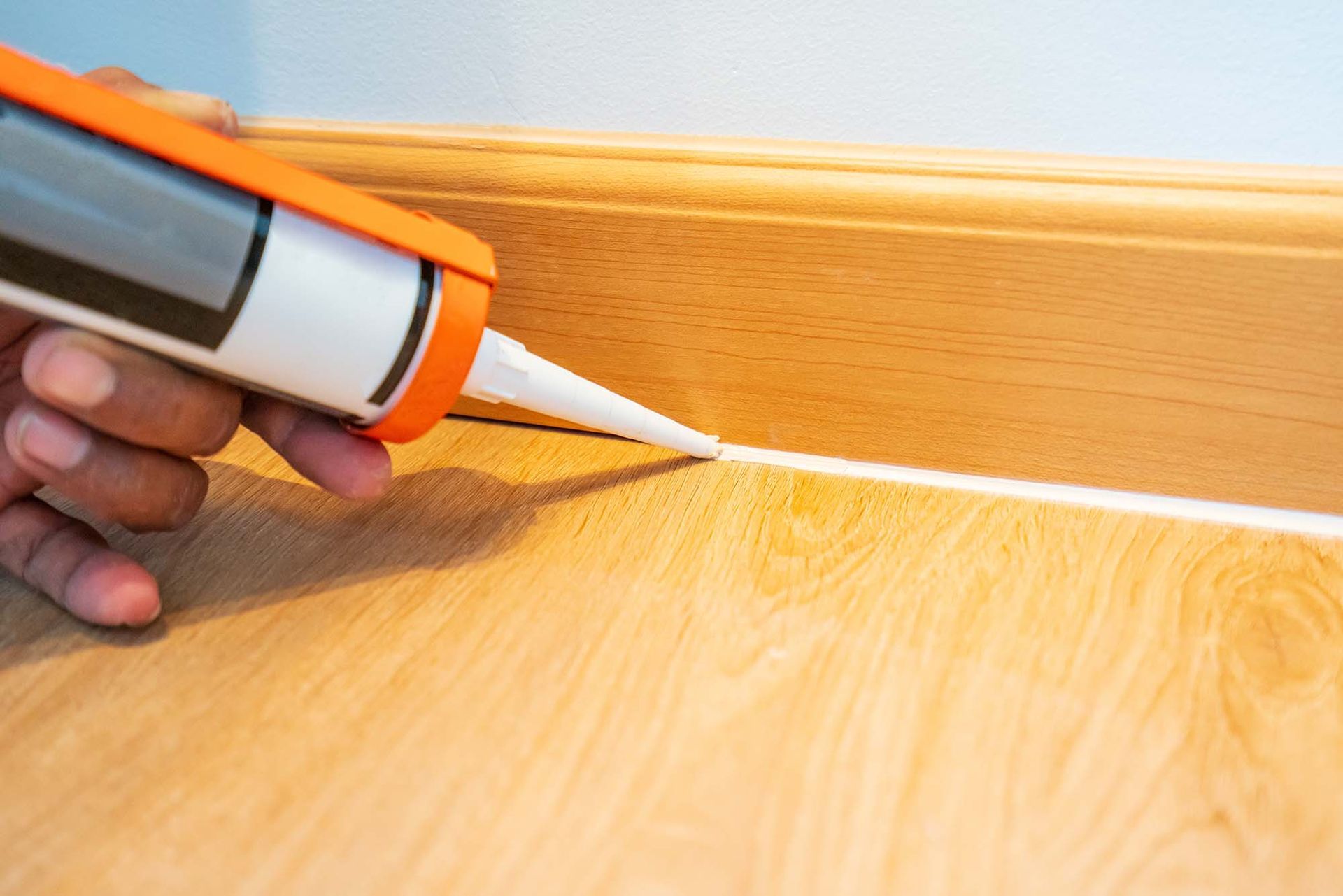 Hand applying white caulk along a wooden floorboard and wall trim with a caulking gun