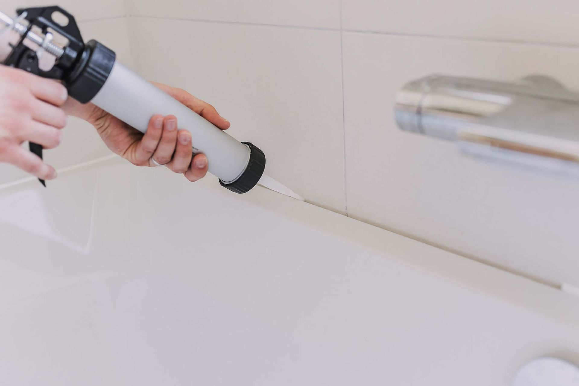 Hand applying caulk along the edge of a white bathtub with a caulking gun