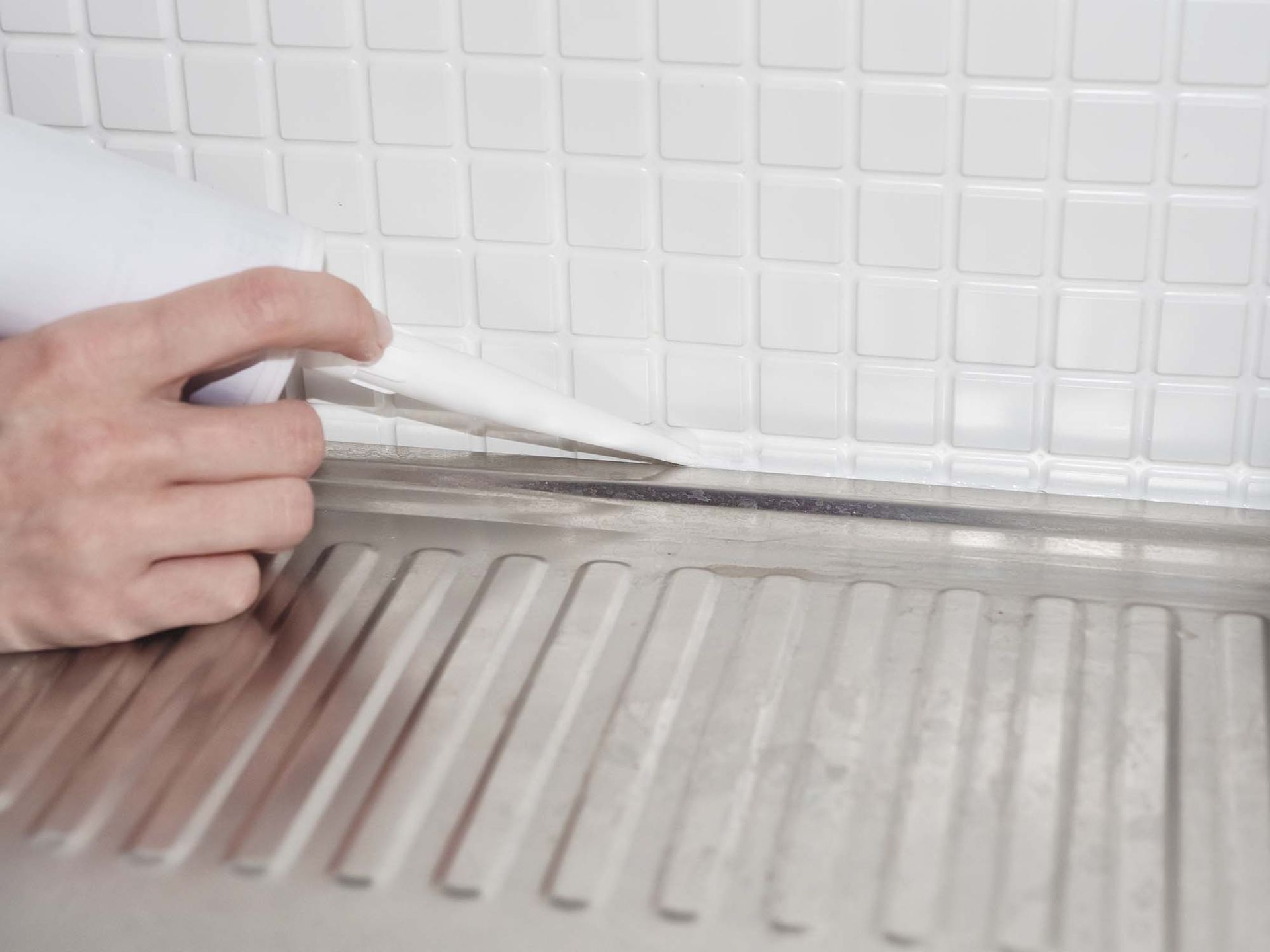 Hand applying white caulk along a backsplash seam above a stainless steel sink