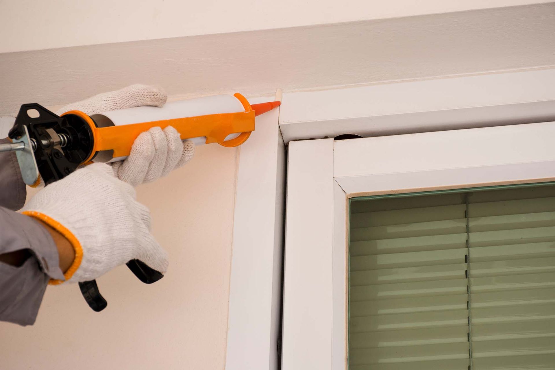 Gloved hands apply orange caulk to the trim above a window.