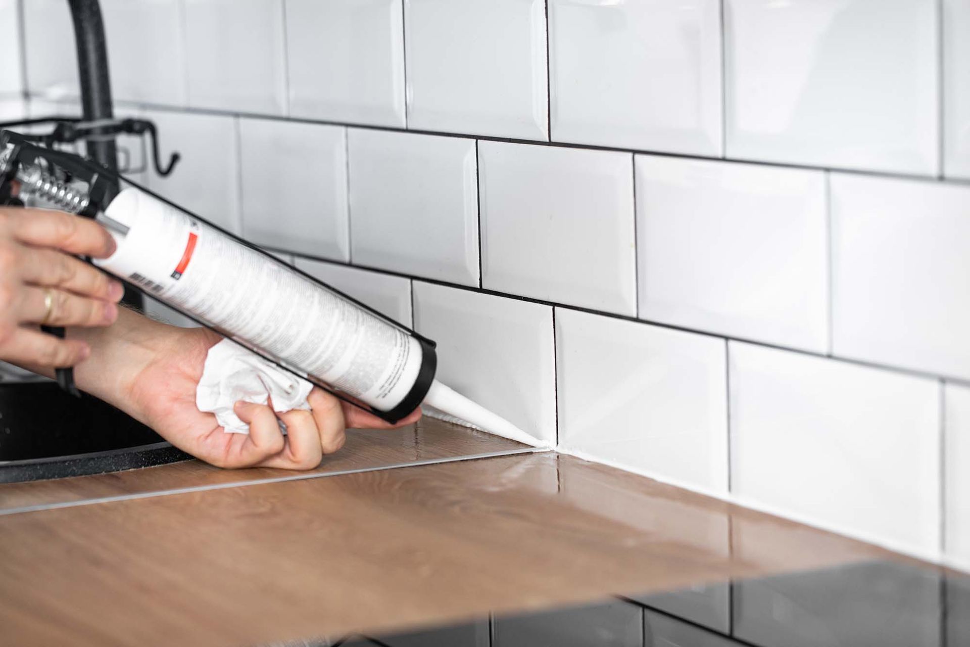 Hands caulking a kitchen backsplash along a white tiled countertop