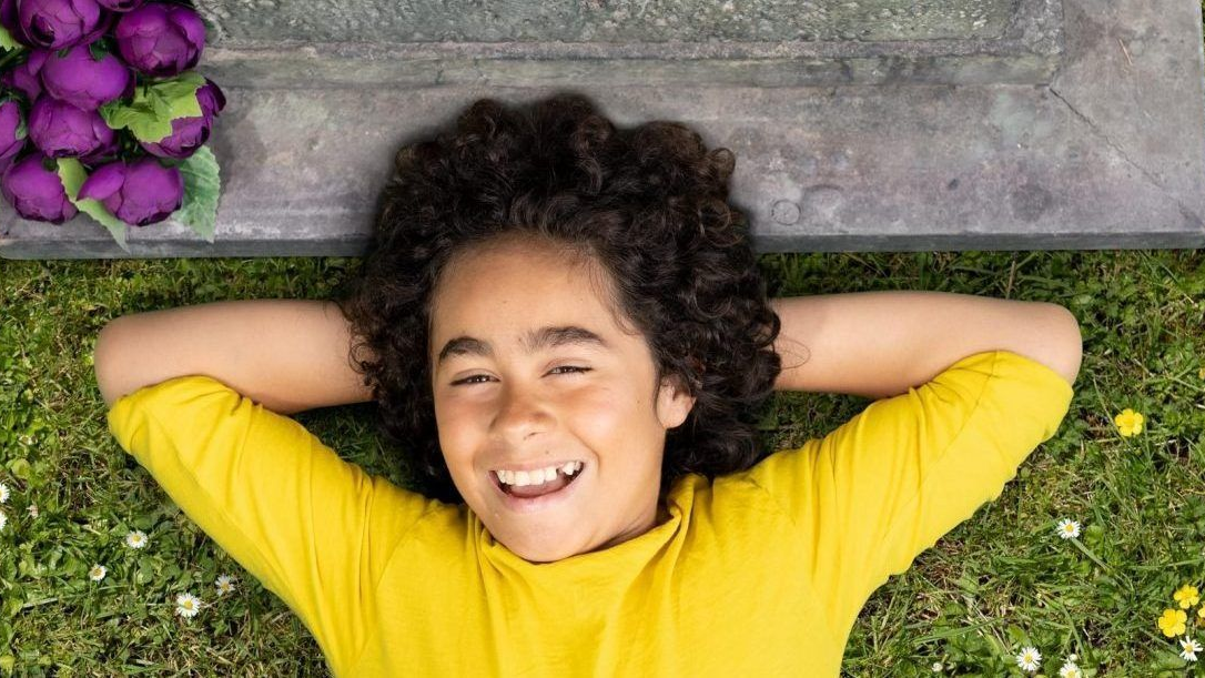 Smiling person lying on grass with hands behind head, wearing a yellow shirt, beside purple flowers.