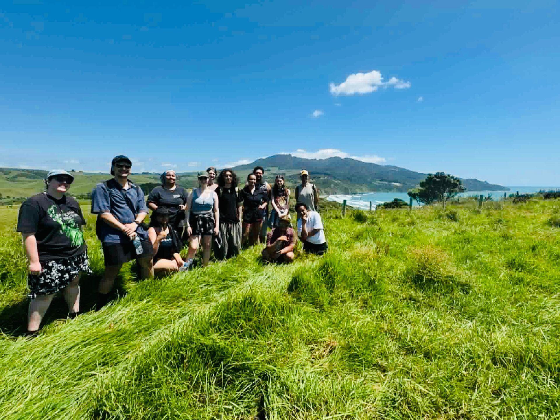 Hikers posing on a grassy hillside with ocean and mountains in the background under a blue sky