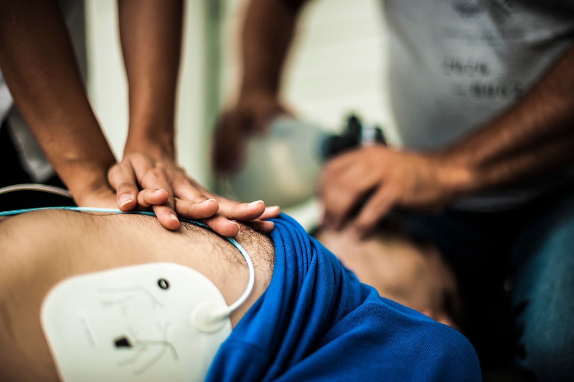 Hands performing CPR on a person's chest, defibrillator pads attached. Another person assists.