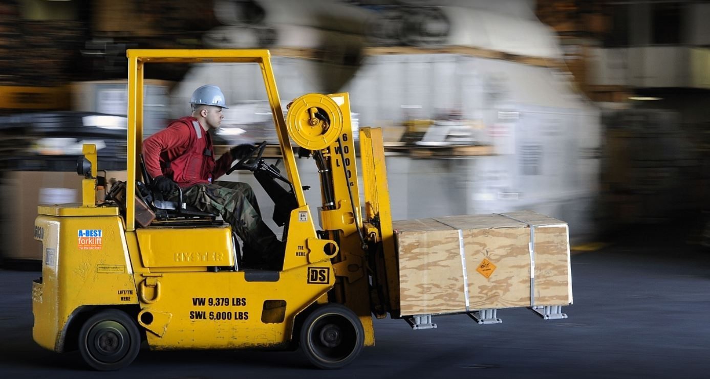 Man operating a forklift, carrying a pallet with a large cardboard box inside a warehouse.