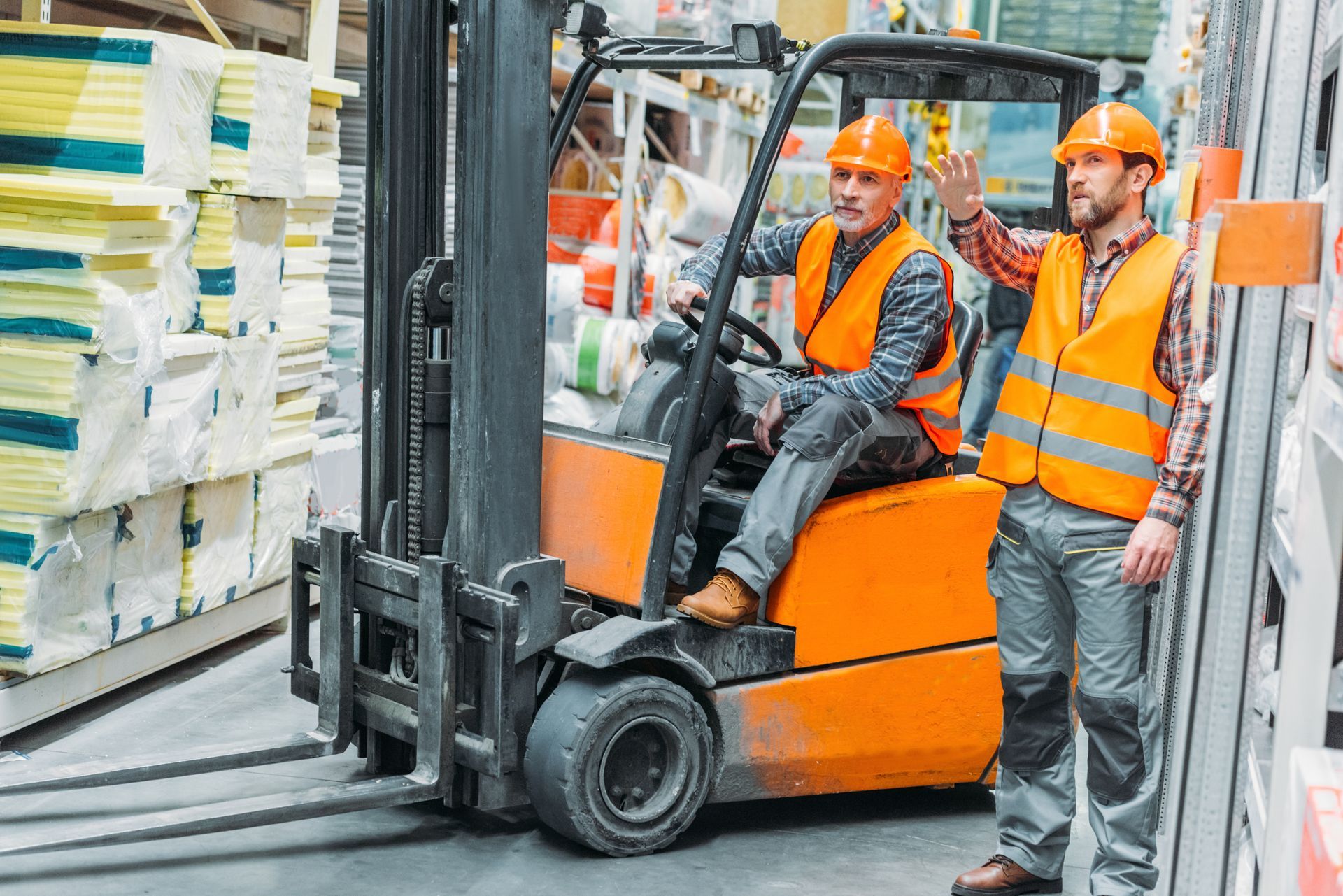 Two workers in orange safety vests and hard hats on a forklift in a warehouse aisle