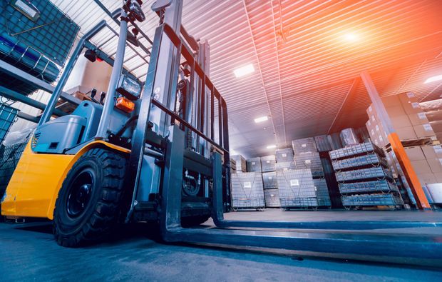 Forklift in a warehouse, moving boxes. Sunlight shines on a busy storage area.
