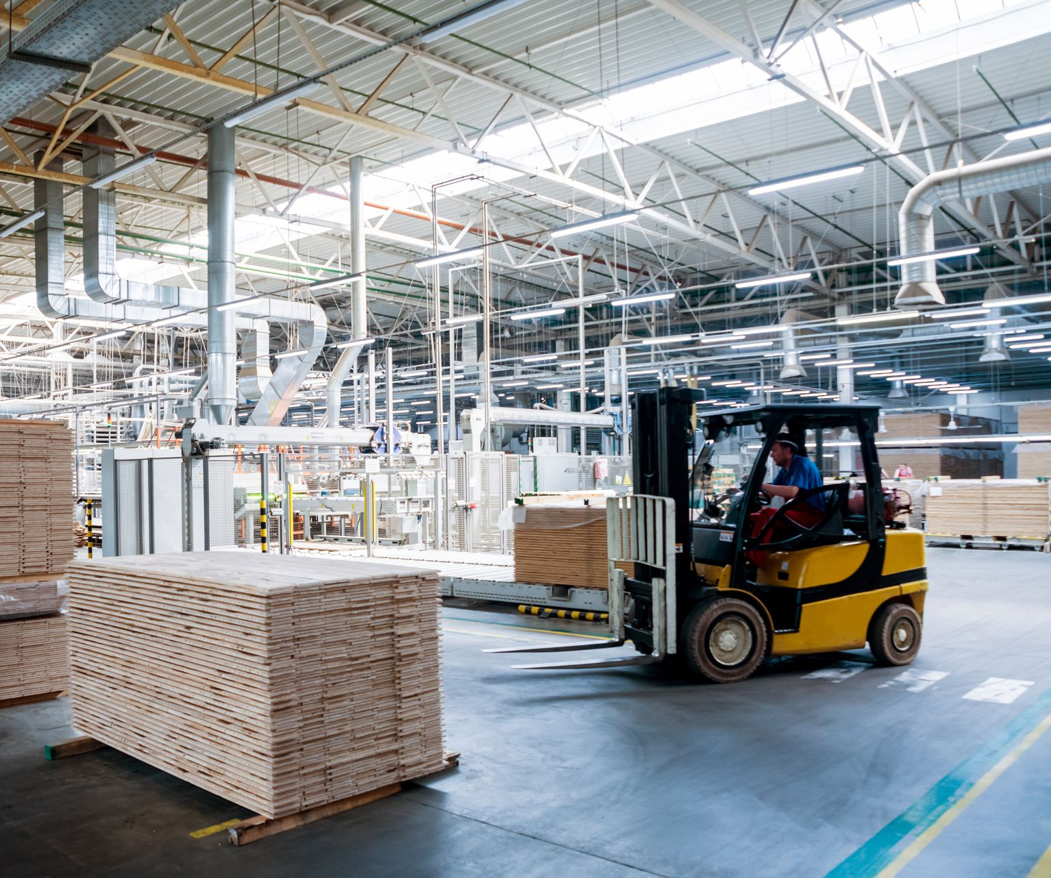 Yellow forklift in a warehouse moves stacks of lumber; a person drives. Yellow forklift in a warehouse moves stacks of lumber; a person drives.