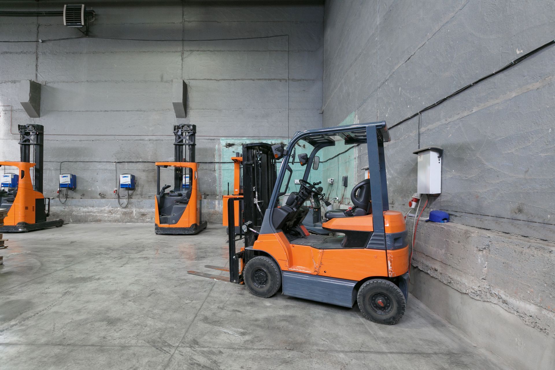 An orange forklift in a warehouse. Other forklifts are visible in the background against a concrete wall. An orange forklift in a warehouse. Other forklifts are visible in the background against a concrete wall.