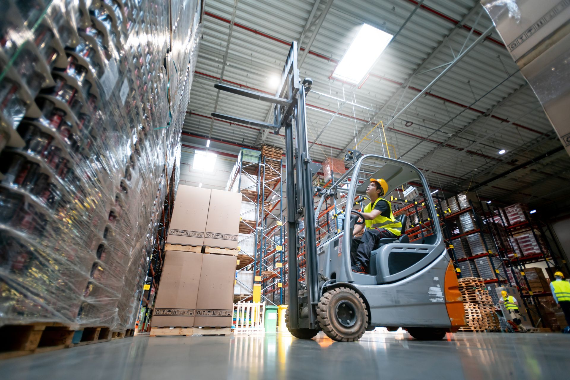 Forklift operator in a warehouse lifting a pallet of boxes. Shelves of goods in background. Forklift operator in a warehouse lifting a pallet of boxes. Shelves of goods in background.