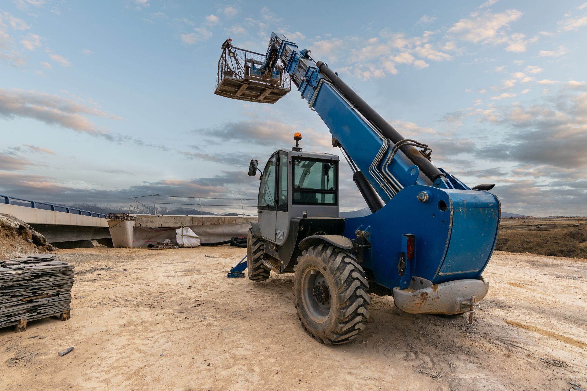 Blue telehandler on a dirt lot with raised bucket under a cloudy sky. Blue telehandler on a dirt lot with raised bucket under a cloudy sky.