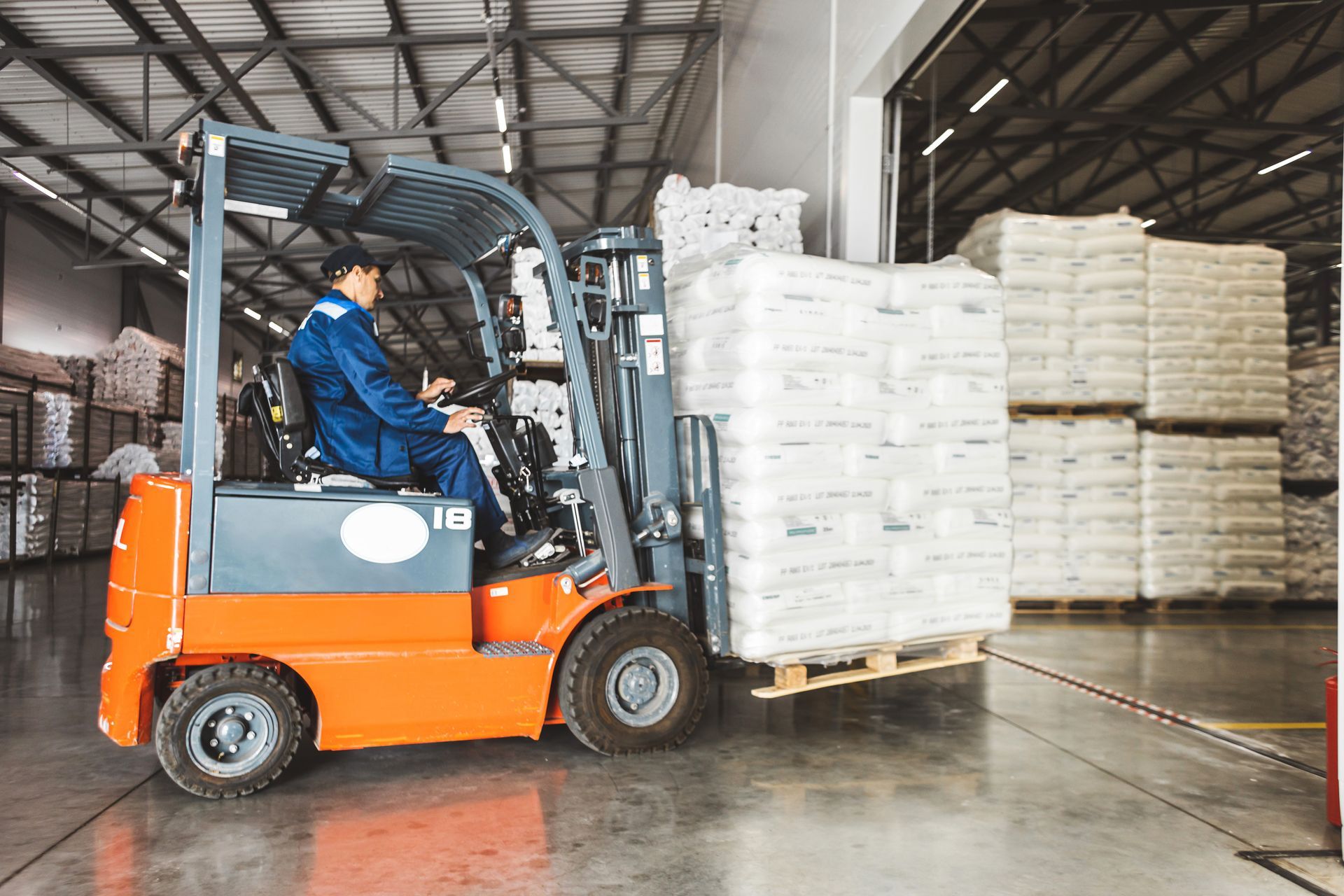 Forklift operator in a warehouse, moving a pallet of wrapped goods. Orange forklift, blue uniform. Forklift operator in a warehouse, moving a pallet of wrapped goods. Orange forklift, blue uniform.