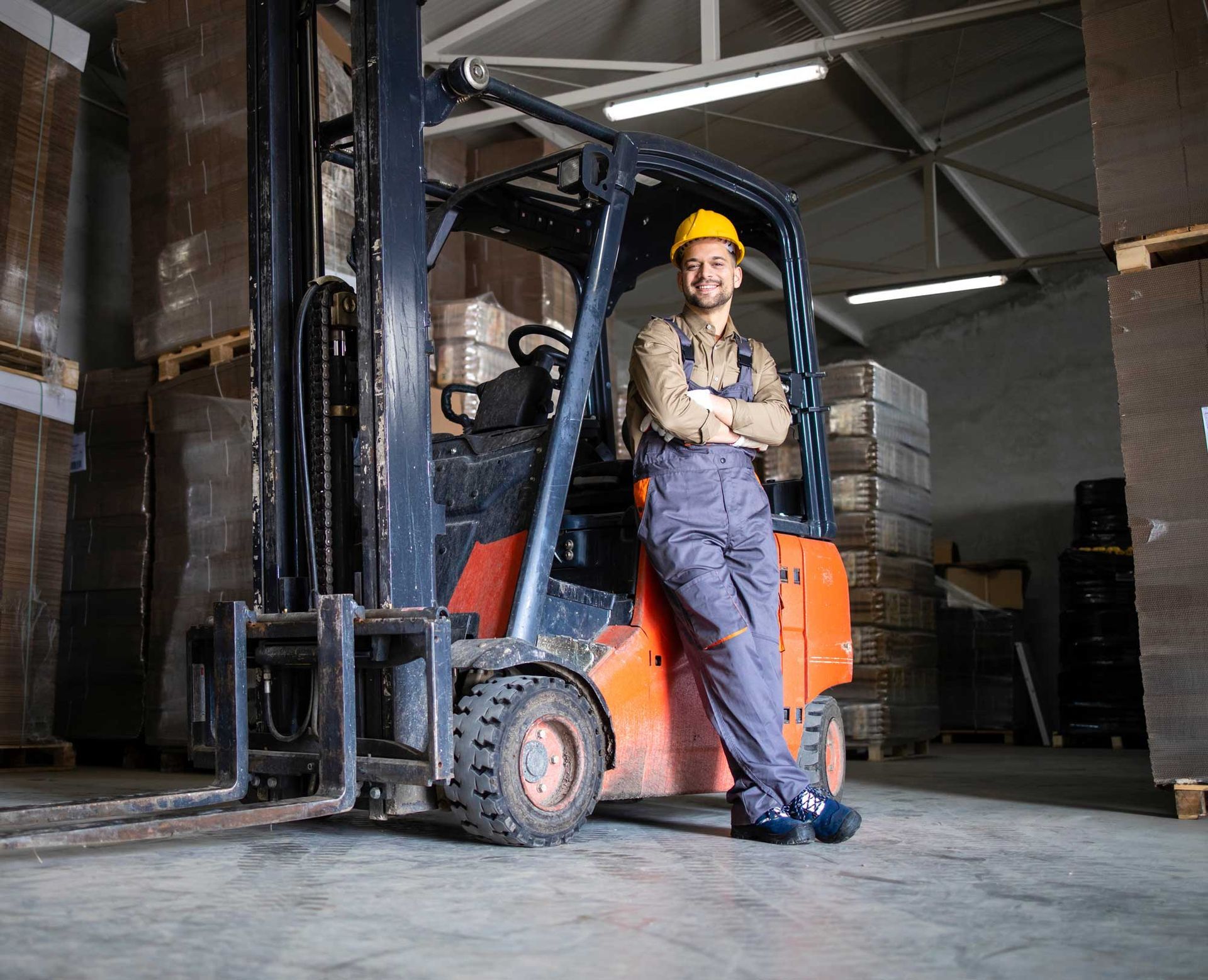 Warehouse worker leaning on forklift, smiling, wearing a hard hat, and coveralls. Inside a warehouse.
