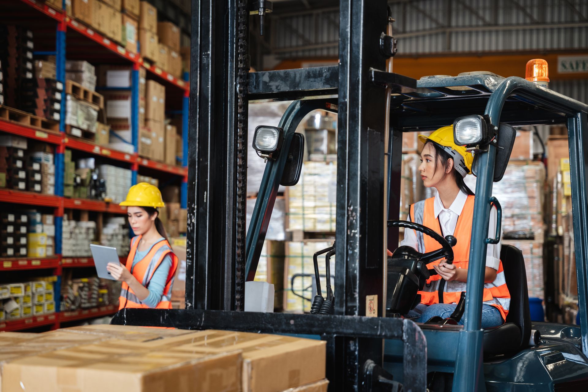 Two women in a warehouse: one driving a forklift, the other using a tablet near shelves of boxes. Two women in a warehouse: one driving a forklift, the other using a tablet near shelves of boxes.