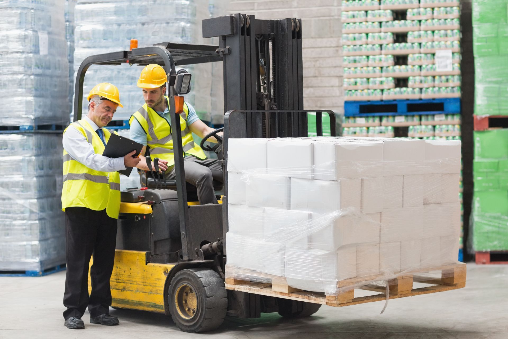 Two workers in a warehouse: One operating a forklift, the other reviewing a clipboard. Two workers in a warehouse: One operating a forklift, the other reviewing a clipboard.